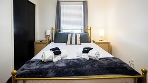 Een opgemaakt bed met handdoeken, een raam met zonwering, bedlampjes en een deur.