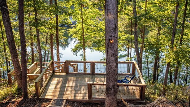 A wooden deck with a hammock and seating area overlooking a lake, surrounded by trees.
