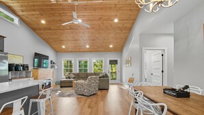 A spacious living area with a wooden ceiling, a dining table with white chairs, a sofa, and a kitchen area with a refrigerator and coffee machine.