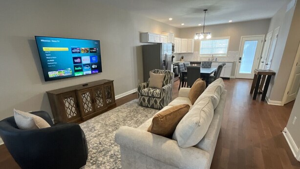 Una sala moderna con un sofá gris, un sillón oscuro y un otoman estampado. Un televisor de pantalla plana está montado en la pared mostrando la interfaz de un servicio de streaming. La habitación cuenta con un mueble de madera y una alfombra con diseño.