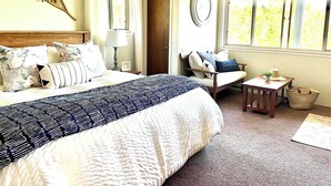 Un dormitorio con una cama grande, una silla, una mesita y una ventana con vistas a árboles.