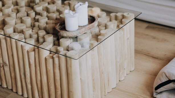 A room with a wooden paneled wall, a white sofa with grey cushions, and a glass coffee table with wooden blocks.