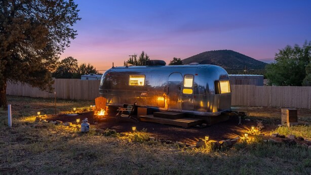 A stationary trailer with a small attached structure, possibly a kitchen or bathroom, in a rural setting with a wooden fence and trees. - Starlight Streamer - Cozy Airstream Escape in Williams, AZ (Williams)