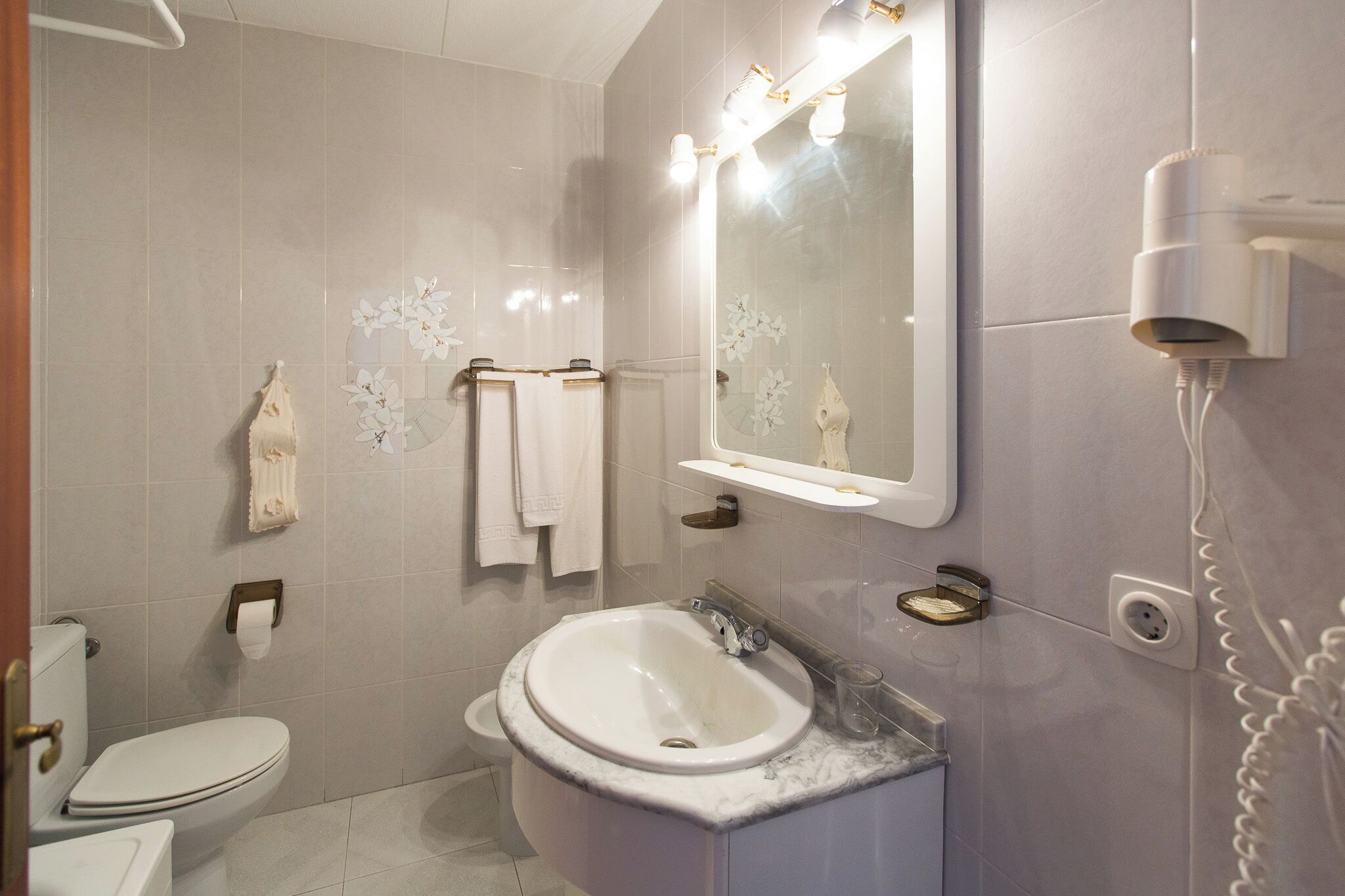 A bathroom with a white toilet, a sink with a marble countertop, a mirror with lights, and a hairdryer mounted on the wall.