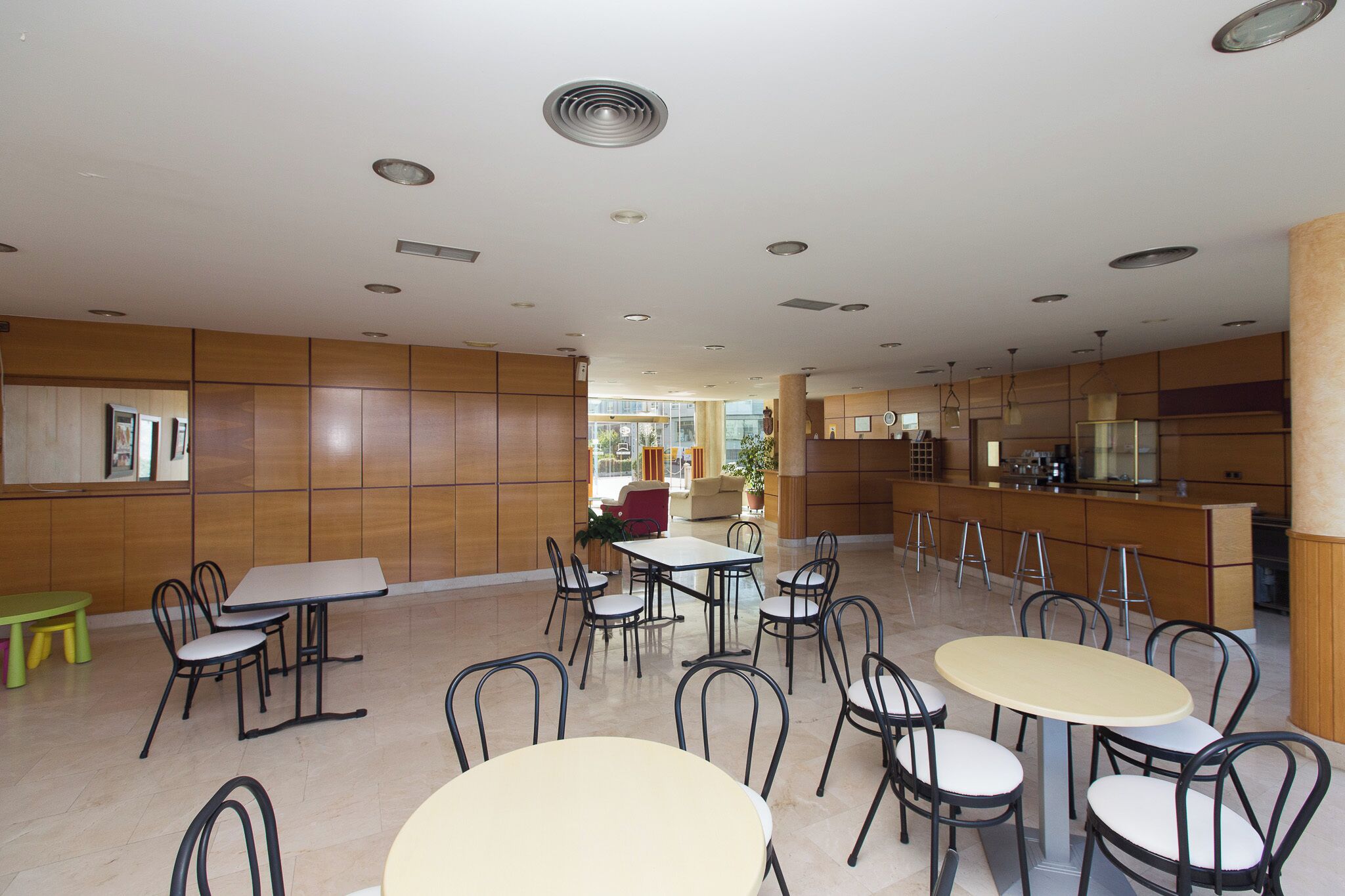 A spacious dining area with multiple tables and chairs, wooden paneling on the walls, and a kitchen area in the background.