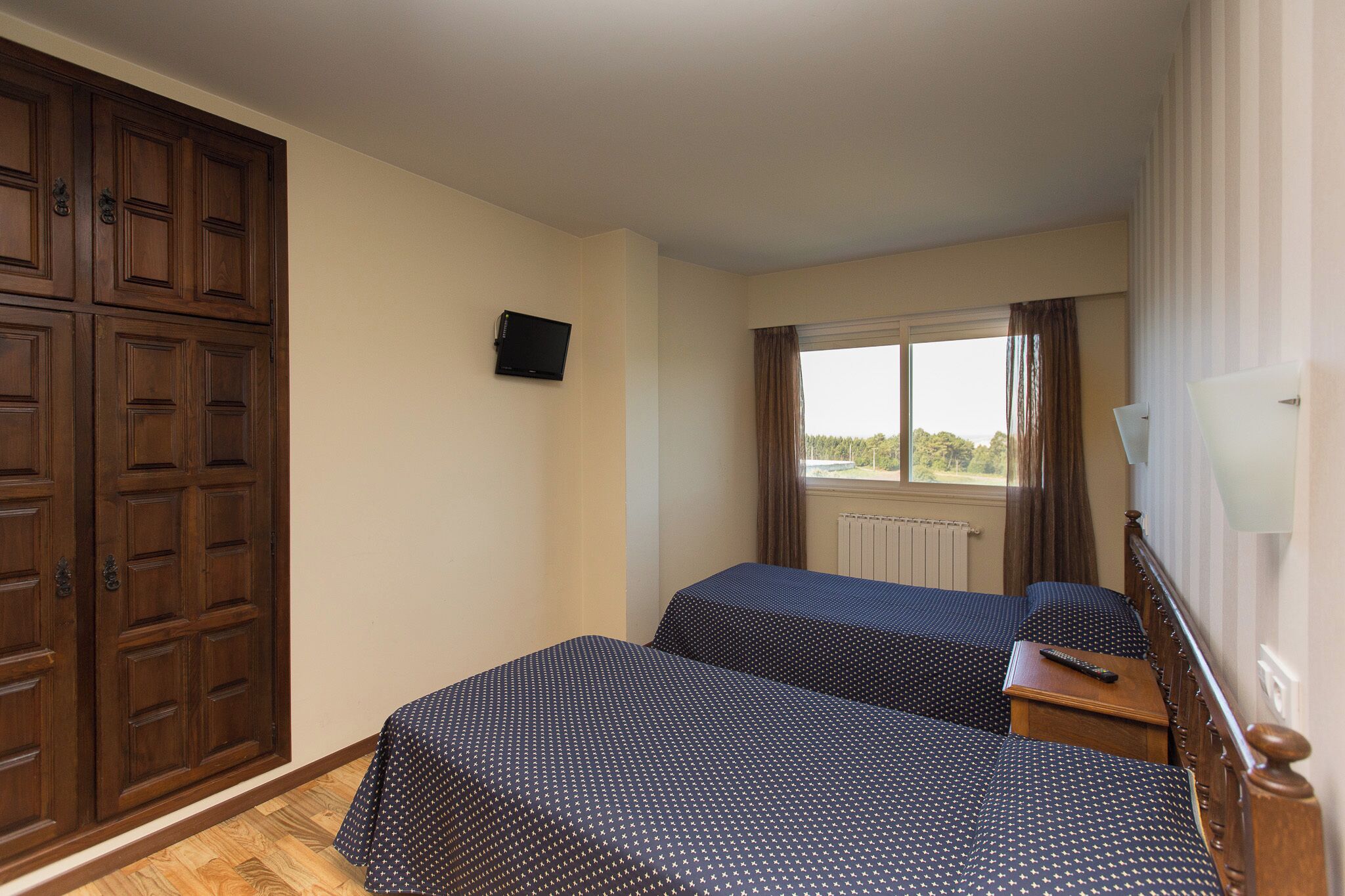 Ein Zimmer mit zwei Betten, einem Fernseher an der Wand, einem Fenster mit Vorhängen und einem Holzschrank.