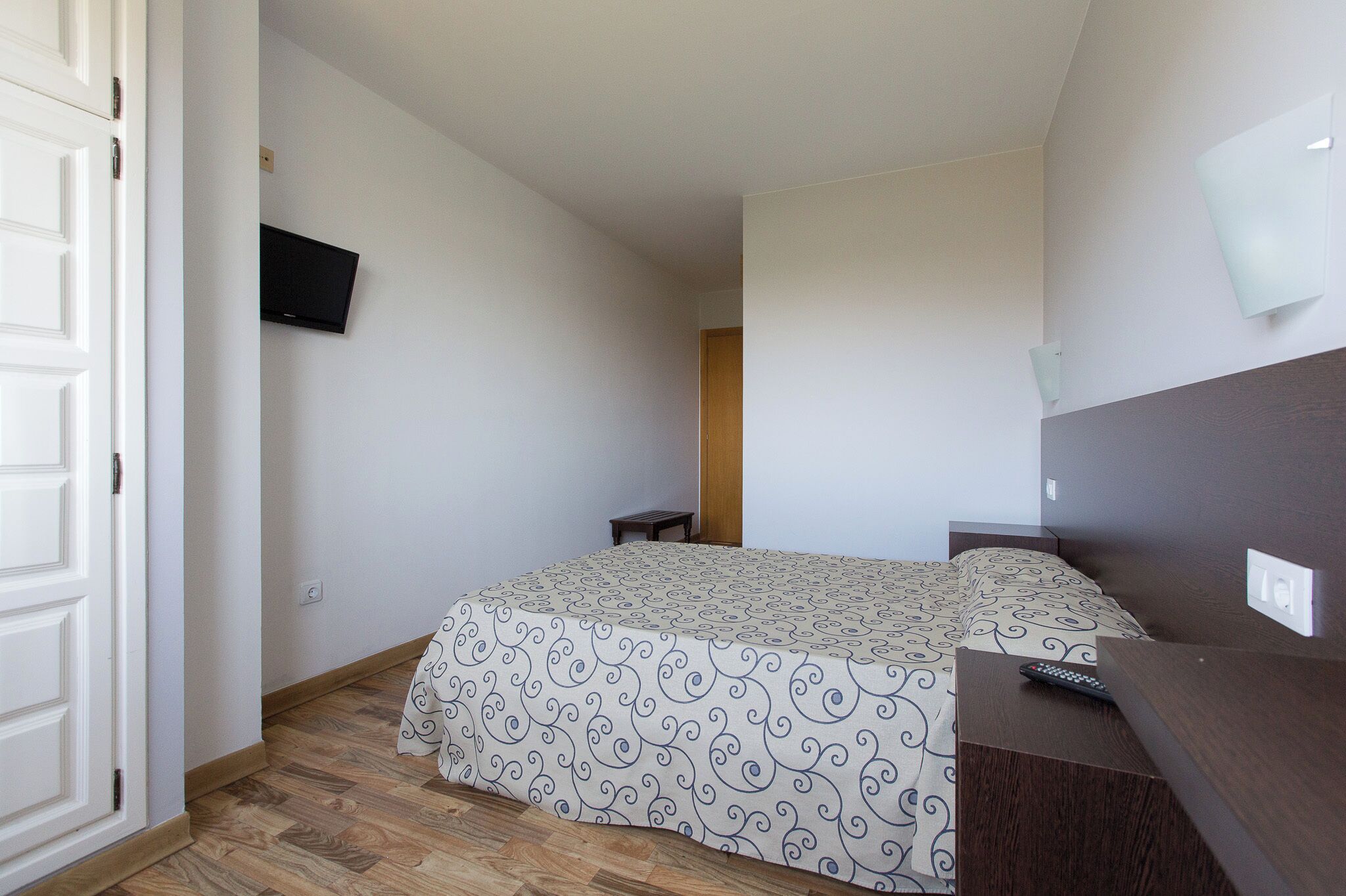 Une chambre à coucher avec un lit, un téléviseur fixé au mur, une petite table et un plancher en bois.