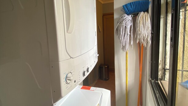 Een wasruimte met een wasmachine en droger, een raam met uitzicht, en een bezem die tegen de muur staat.