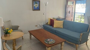 Een woonkamer met een blauwe bank, een houten salontafel, een rieten fauteuil en een bijzettafeltje met een bloemenvaas.