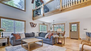 A spacious living area with a large sofa, a coffee table, and a dining table with chairs. There is a balcony with a railing and a view of trees. - Hunt Hideaway, 4bd 3ba sleeps 12, hot tub, 85” TV, arcades! (Albrightsville)