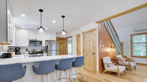 A modern kitchen with white cabinets, a central island with bar stools, and a wooden staircase. - Hunt Hideaway, 4bd 3ba sleeps 12, hot tub, 85” TV, arcades! (Albrightsville)