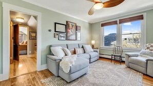 A living room with a grey sofa, a wooden ceiling fan, and framed pictures on the wall. - BozemanGetaways - Charming Condo Steps to Main (Bozeman)