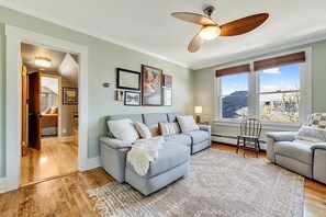 A living room with a grey sofa, a wooden ceiling fan, and framed pictures on the wall. - BozemanGetaways - Charming Condo Steps to Main (Bozeman)