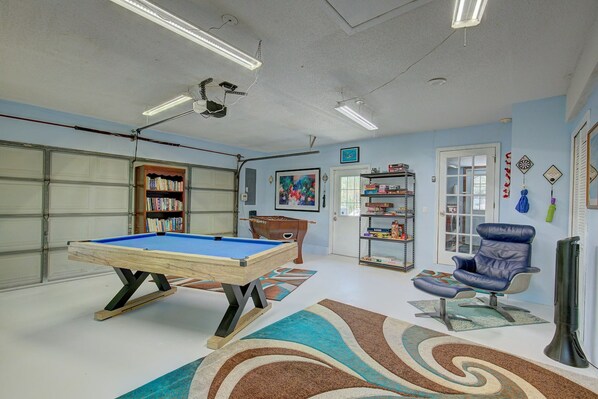 Una habitación con mesa de billar, un sillón azul, una estantería y una puerta que da a un garaje.