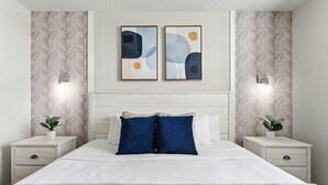 A bedroom with a bed, two bedside tables, a nightstand, a wall-mounted light, and a herringbone-patterned wallpaper.