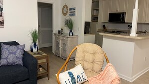 Ein Wohnzimmer mit einem dunkelblauen Sofa, einem Sessel mit Kissen mit dem Schriftzug 'HOME', einem kleinen Beistelltisch mit Pflanze und einem Wandschild mit der Aufschrift 'Home'.