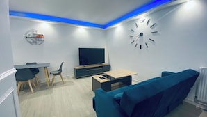 Ein modernes Wohnzimmer mit einem blauen Sofa, einem Fernseher, einem Holztisch und einem Essbereich mit Stühlen und einem kleinen Tisch.