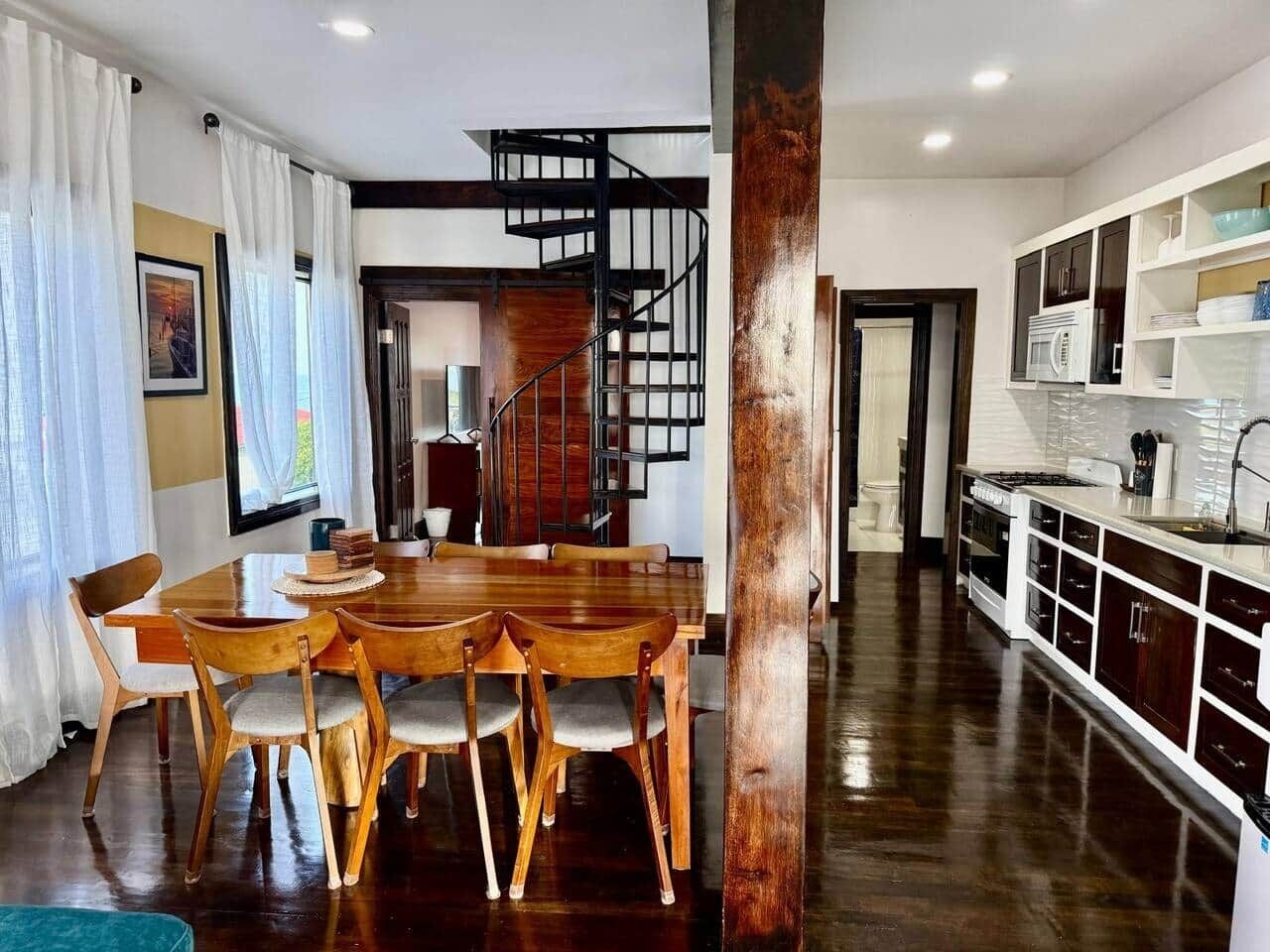 Un vaste espace dédié à la salle à manger, avec une table et des chaises en bois, un escalier et un coin cuisine doté de meubles blancs et d’une cuisinière.