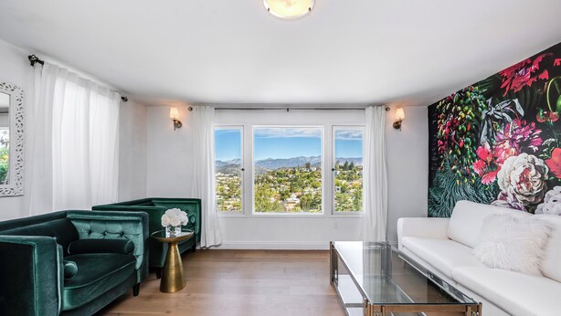 A modern living room with a green sofa, a white sofa, and a glass coffee table. There are three windows with a view of a cityscape and mountains. - Stunning 1-bedroom apartment with view (Glendale)