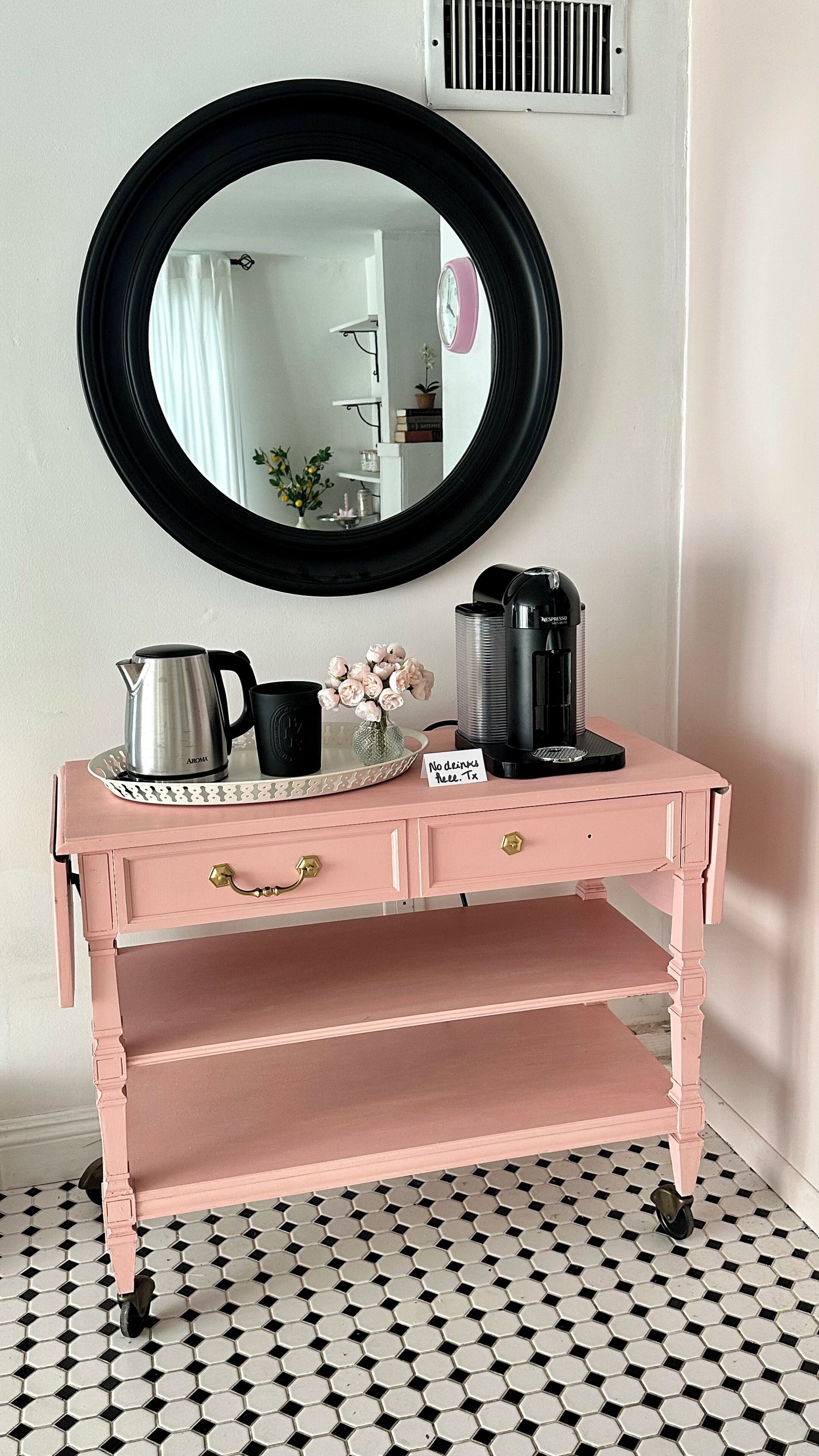 A pink vanity with a round mirror, a tray with a coffee maker, and a sign indicating no drinks allowed.