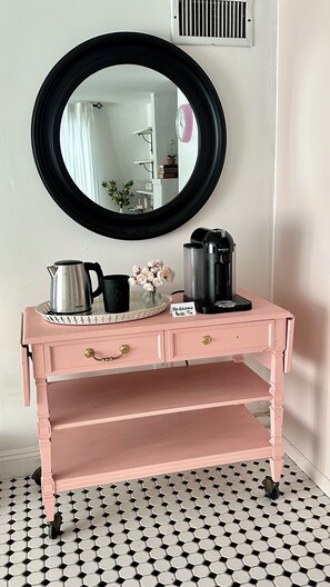 A pink vanity with a round mirror, a tray with a coffee maker, and a sign indicating no drinks allowed.