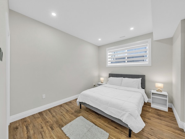 A bedroom with a bed, nightstands, a window with blinds, and a wooden floor.
