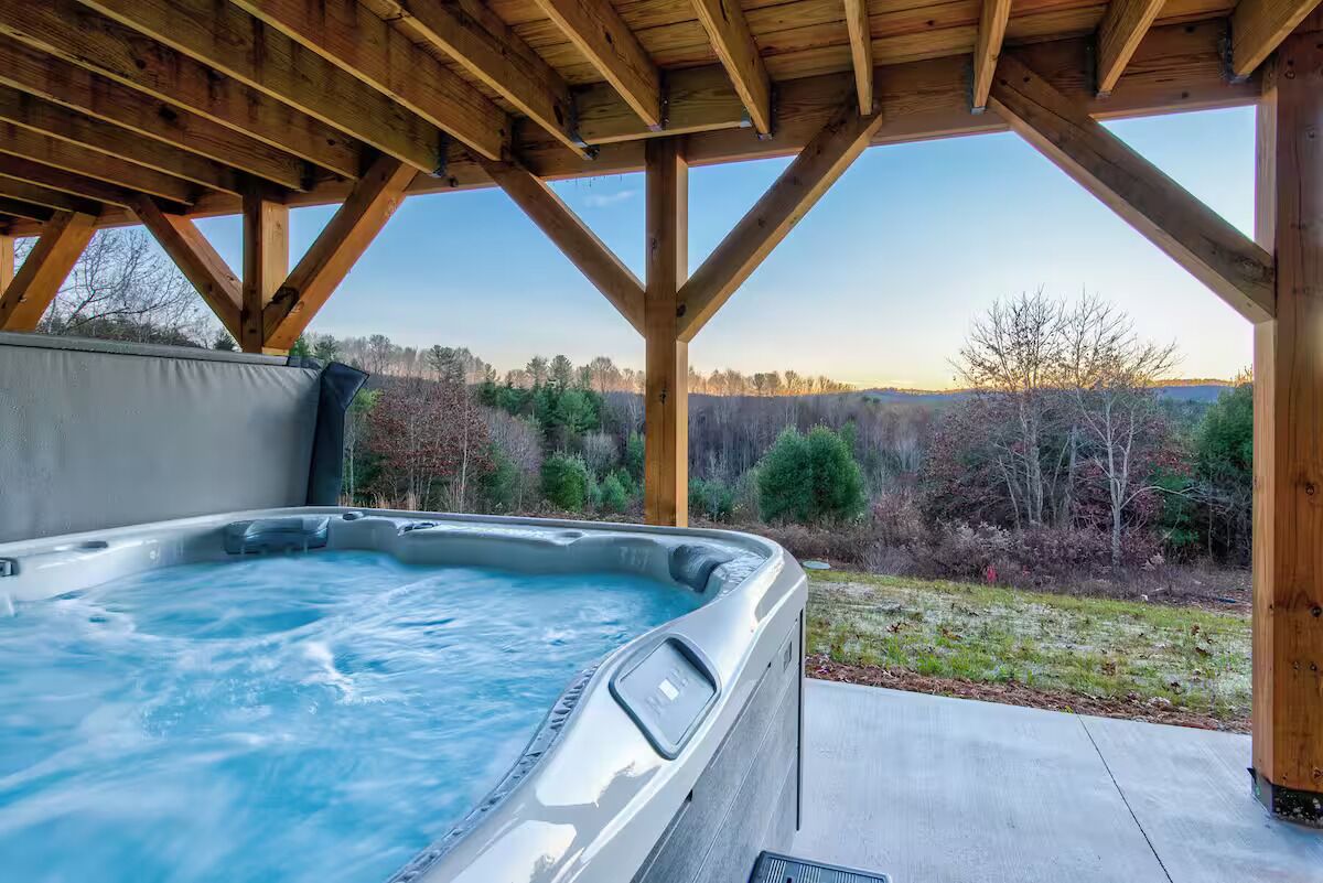 A hot tub under a wooden pergola with a view of a forested landscape.