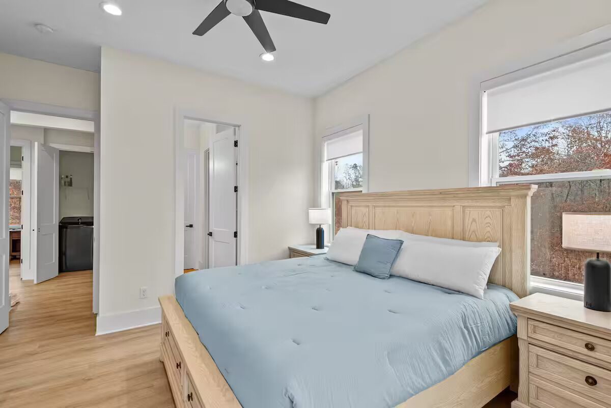 A bedroom with a bed, wooden headboard, bedside tables, a ceiling fan, and a window with a view.
