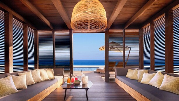Uma área de estar ao ar livre luxuosa, com piso de madeira, sofás acolchoados e uma mesa central com bebidas e petiscos.