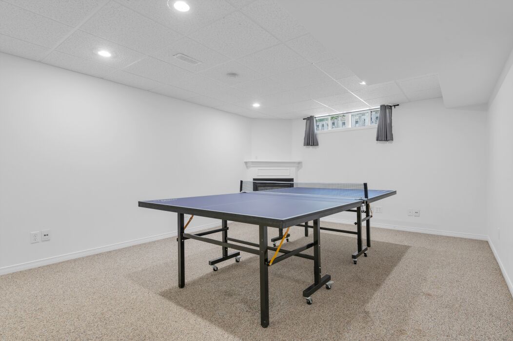 A room with a ping pong table, carpeted floor, white walls, and a fireplace.