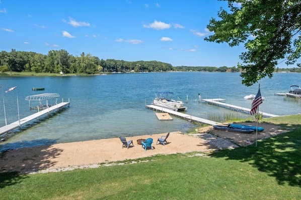 A lakeside dock with boats, a sandy beach with chairs, and an American flag.