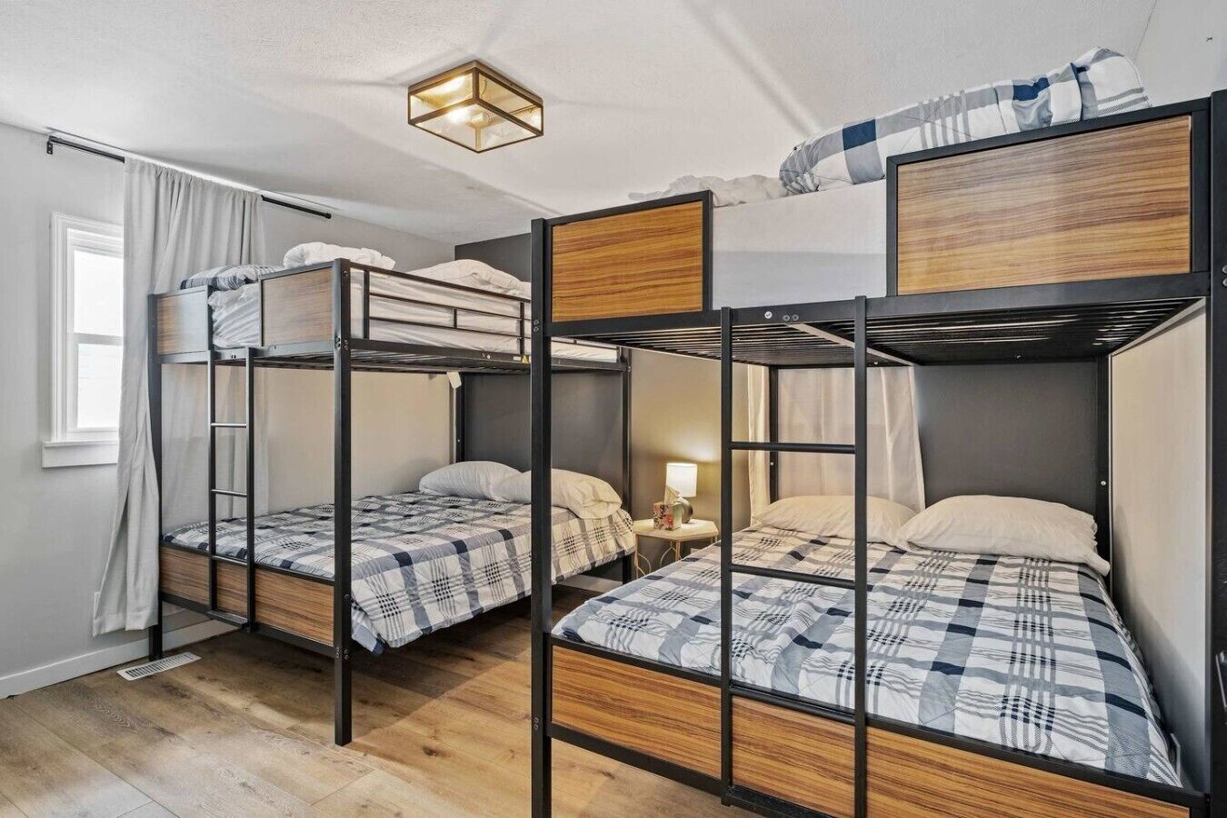 Ein Schlafsaalzimmer mit Etagenbetten, einem Fenster mit Vorhängen, einem Nachttisch mit Lampe und einer Deckenleuchte.