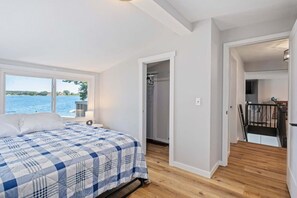Une chambre avec un lit, une fenêtre donnant sur l’eau, un dressing et un escalier menant à une autre pièce.