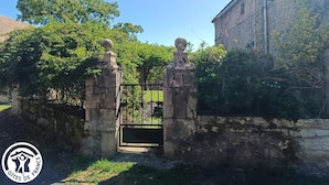 A stone gate with two pillars and a metal gate, surrounded by greenery and a building with a stone facade.