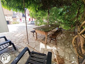 Un patio al aire libre con una mesa y bancos de madera, rodeado de piso de piedra y follaje.