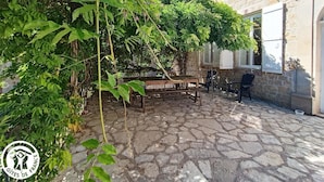 A stone-paved courtyard with a wooden bench and chairs, surrounded by lush greenery and a stone building with windows.