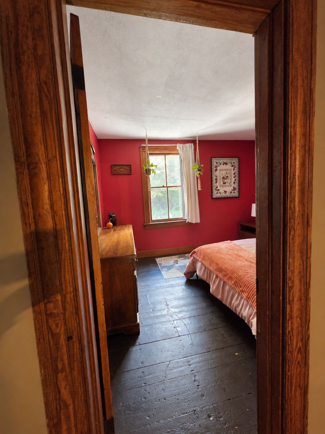Een slaapkamer met een houten vloer, een bed met een roze sprei, een raam met gordijnen en een houten dressoir.