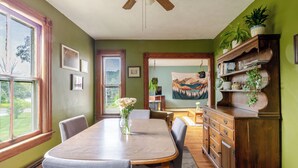 A dining room with a wooden table, chairs, and a vase of flowers. There is a window with a view of greenery outside.