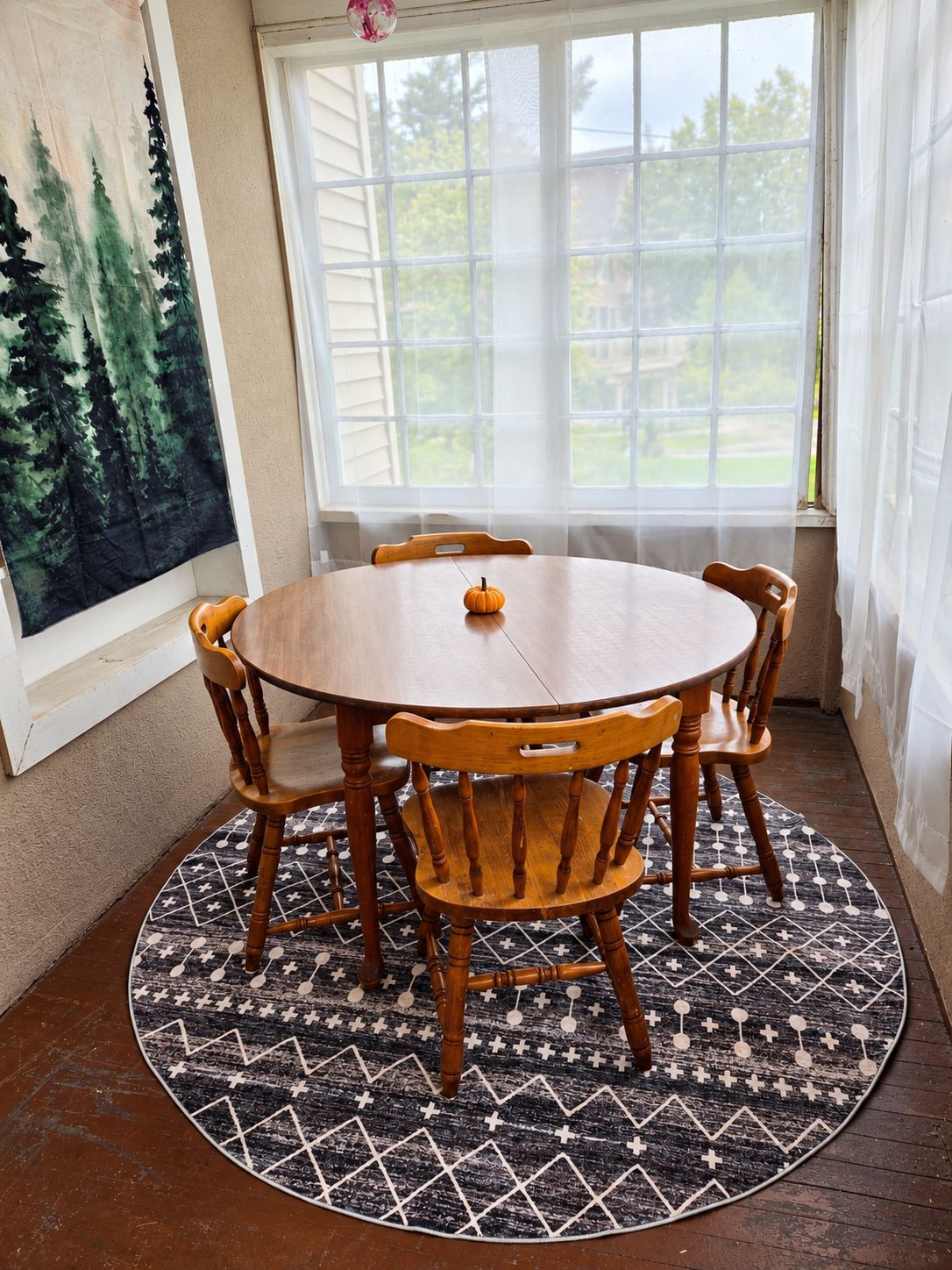 Una mesa de comedor redonda de madera con seis sillas, un centro de mesa con una calabaza pequeña y una alfombra estampada sobre un piso de madera.