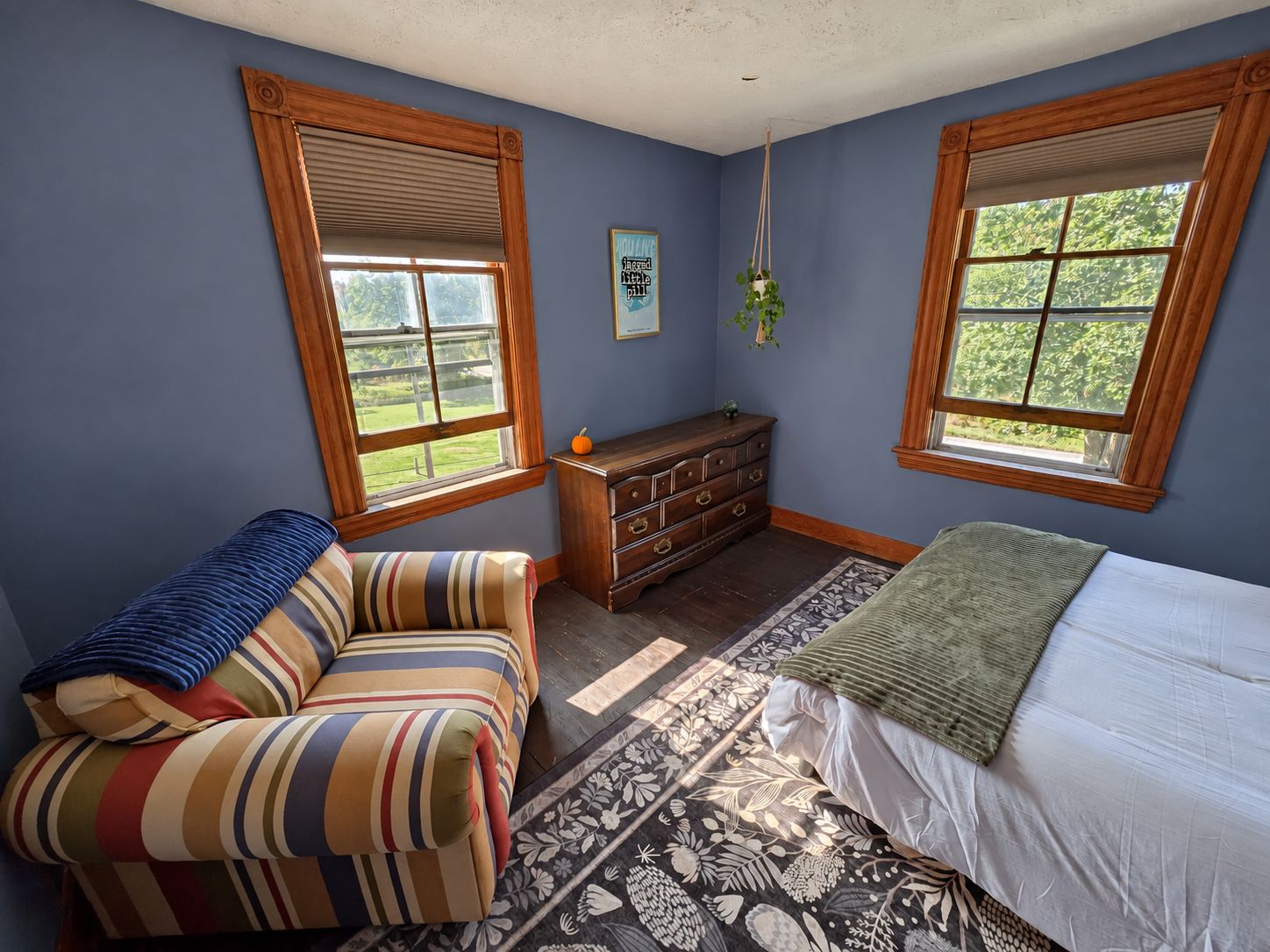 A bedroom with a bed, a striped armchair, a dresser, and two windows with blinds.