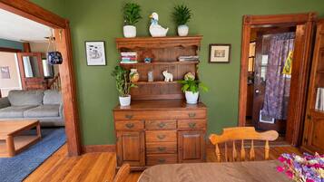 Ein Zimmer mit einem Holzkapstativ, einem Bett, einem Esstisch und einem Sofa. Auf dem Kapstativ stehen Blumentöpfe und auf dem Esstisch eine Vase mit Blumen.