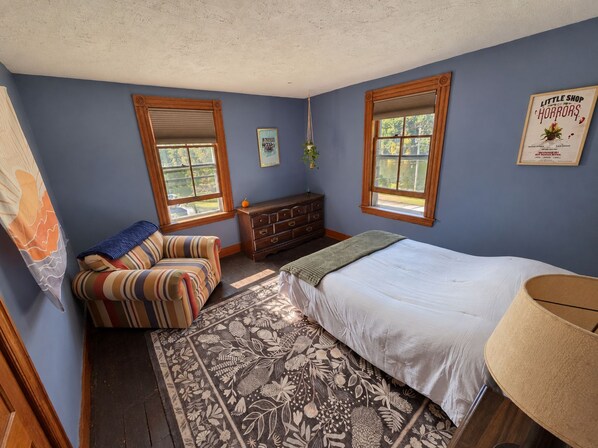 A bedroom with a bed, a dresser, a chair, a window, and a wall with a framed picture.