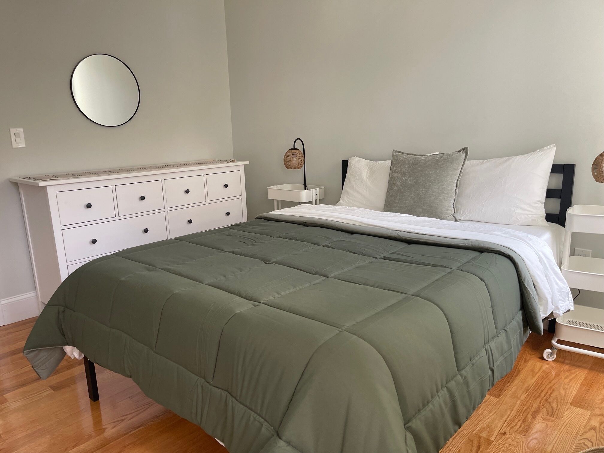 Una cama bien hecha con edredón verde, un aparador blanco con cajones, un espejo redondo en la pared y una mesita de noche con lámpara.