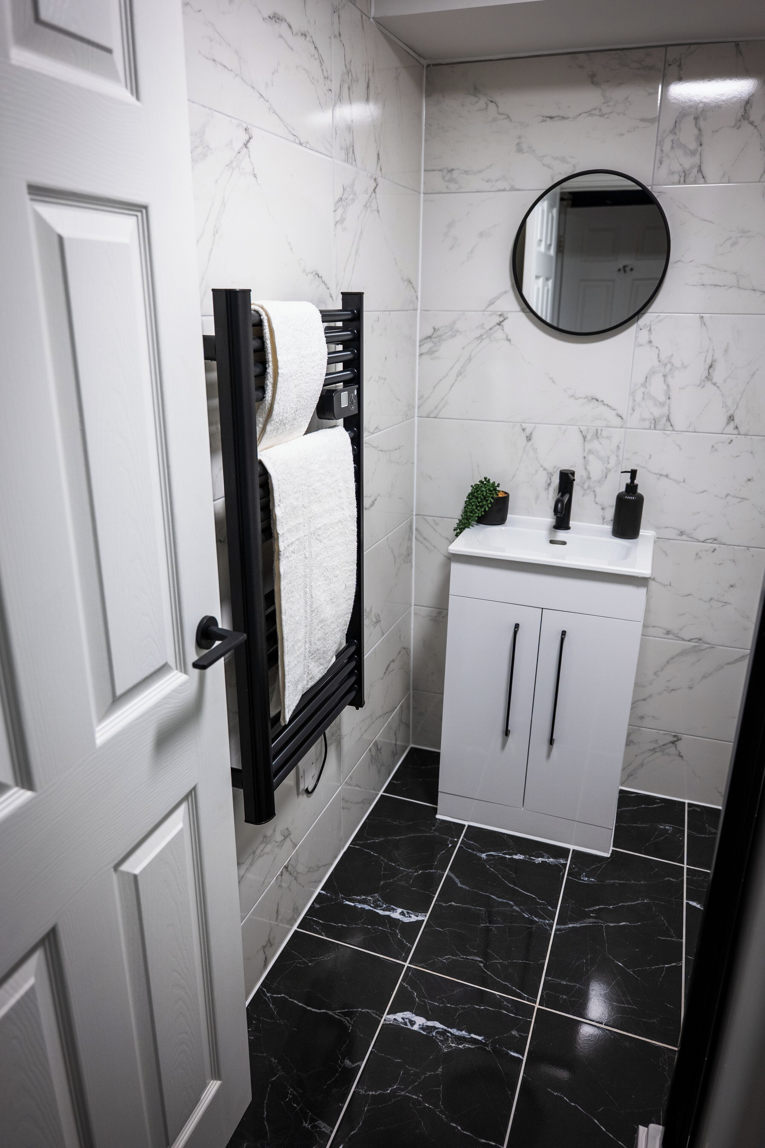 A bathroom with a white door, a towel rack with a white towel, a white cabinet with black handles, a black sink with a mirror above it, and black marble tiles on the floor.