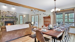 A cozy living room with a fireplace, a dining table set for six, and a view of the outdoors through large windows. - Spa-Like Rustic Retreat In Nature With Glass Barrel Sauna, Hot Tub & Pond! (Albrightsville)