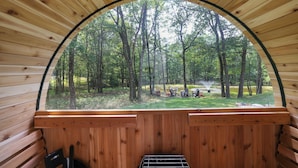 A wooden cabin with a large arched window offering a view of a forested area with outdoor seating and a grill. - Spa-Like Rustic Retreat In Nature With Glass Barrel Sauna, Hot Tub & Pond! (Albrightsville)