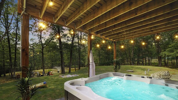 A hot tub with a wooden deck and string lights, surrounded by a grassy area and trees. - Spa-Like Rustic Retreat In Nature With Glass Barrel Sauna, Hot Tub & Pond! (Albrightsville)