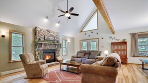 A cozy living room with a stone fireplace, comfortable seating, and a view of the outdoors. - Spa-Like Rustic Retreat In Nature With Glass Barrel Sauna, Hot Tub & Pond! (Albrightsville)