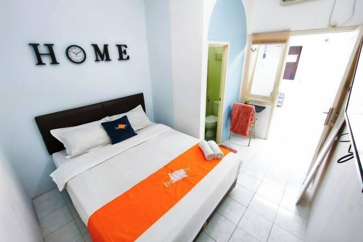 Una habitación de hotel con una cama, un cartel que dice 'HOME' en la pared y un baño visible a través de un arco.