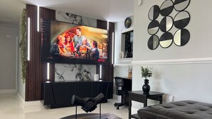 Ein modernes Wohnzimmer mit einem großen Flachbildfernseher an einer Marmorwand, einer schwarzen Ledersofa, einem kleinen runden Tisch mit einer Skulptur und einem dekorativen Wandbild.
